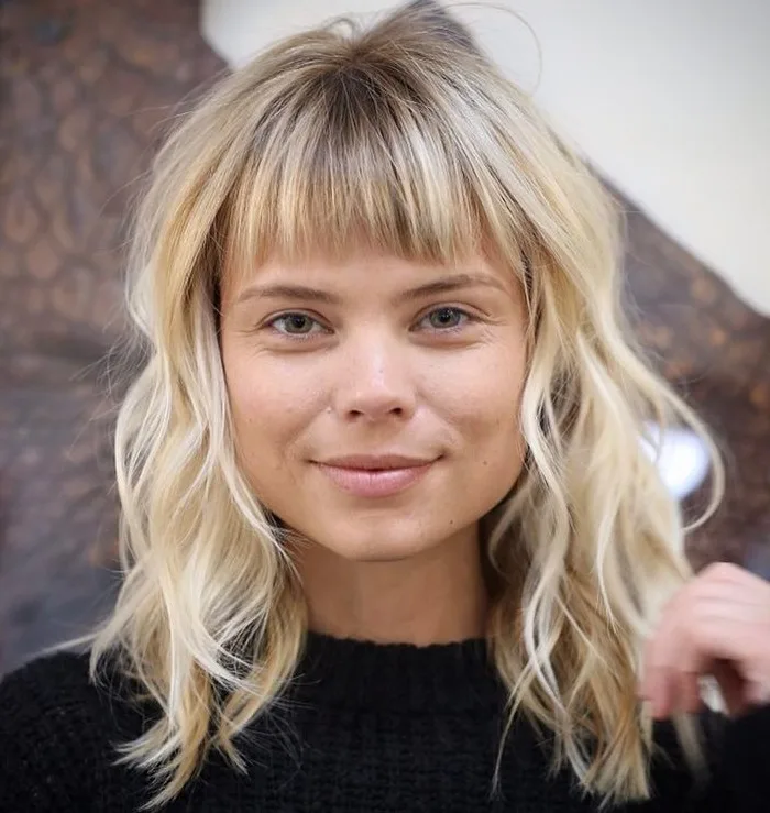Coupe mi-longue blonde ondulée avec frange fournie adoucissant un visage carré
