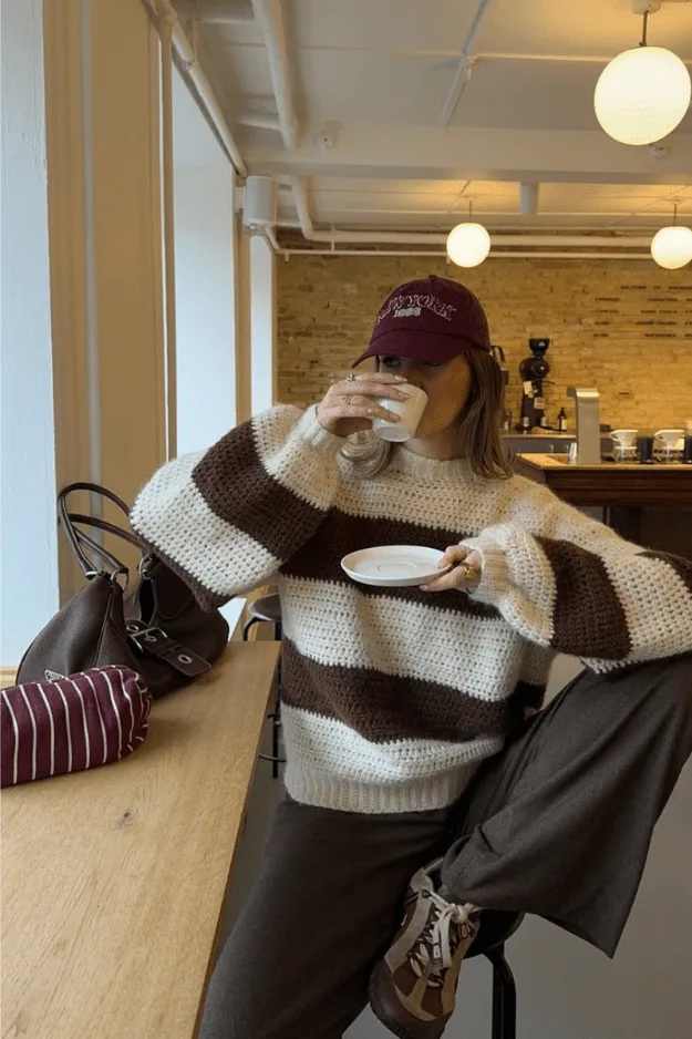 Femme en pull rayé oversize avec pantalon marron, café à la main dans un salon de thé