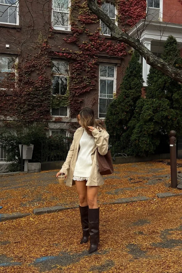 Femme en trench crème avec bottes hautes sous les feuilles d'automne
