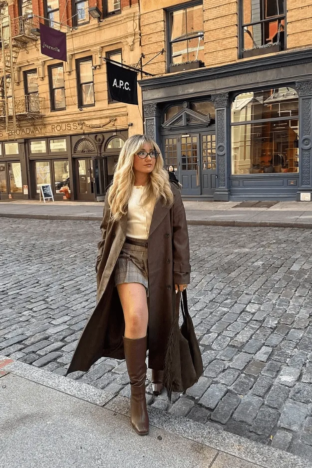 Look trench long chocolat, top crème et mini-jupe à carreaux marron avec bottes