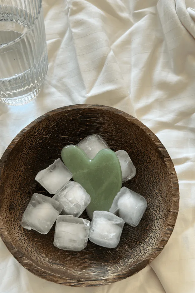 Bol de glaçons avec un outil de gua sha en pierre, sur un lit clair