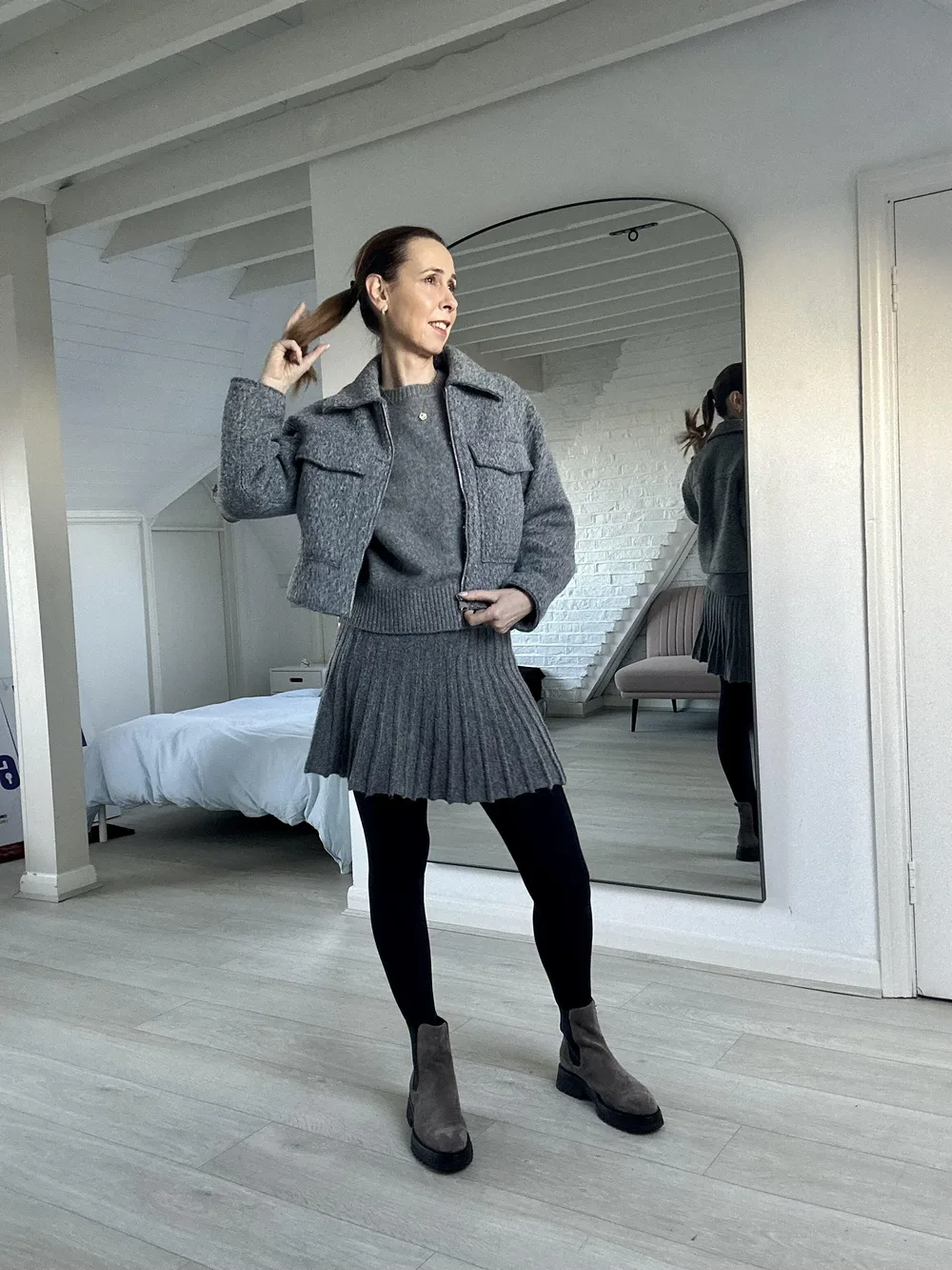 Femme de 60 ans dans une tenue grise ton sur ton avec jupe plissée et veste en maille