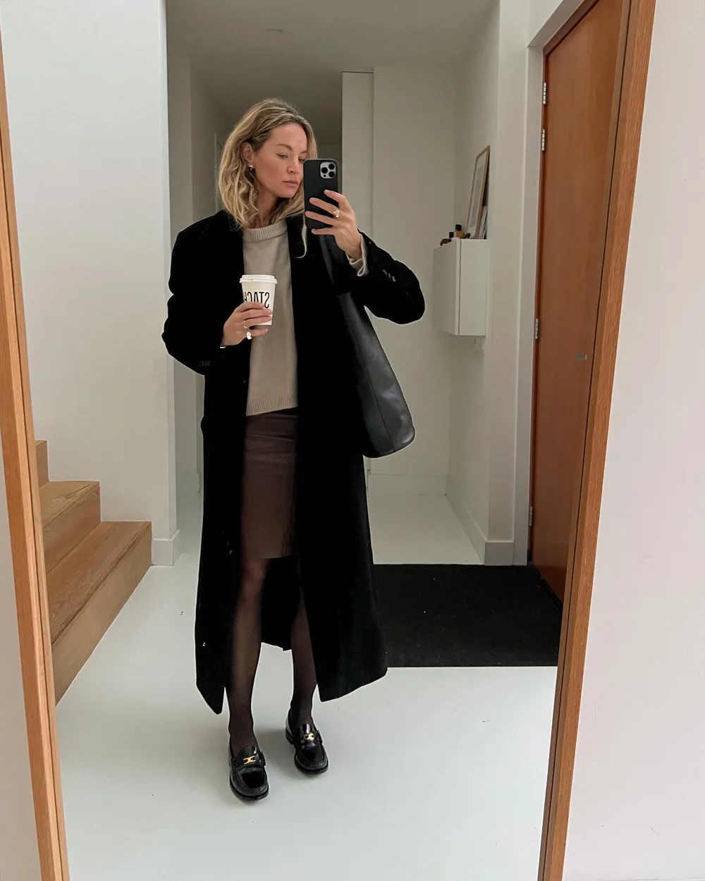 Long manteau noir porté avec une jupe marron, des collants et des mocassins
