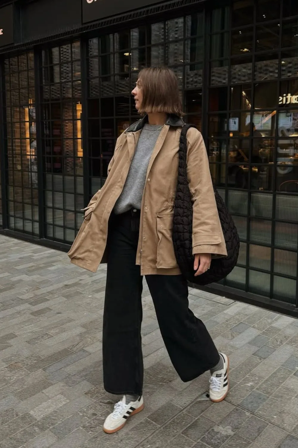 Pull gris porté avec une veste workwear beige, un jean noir large et des baskets