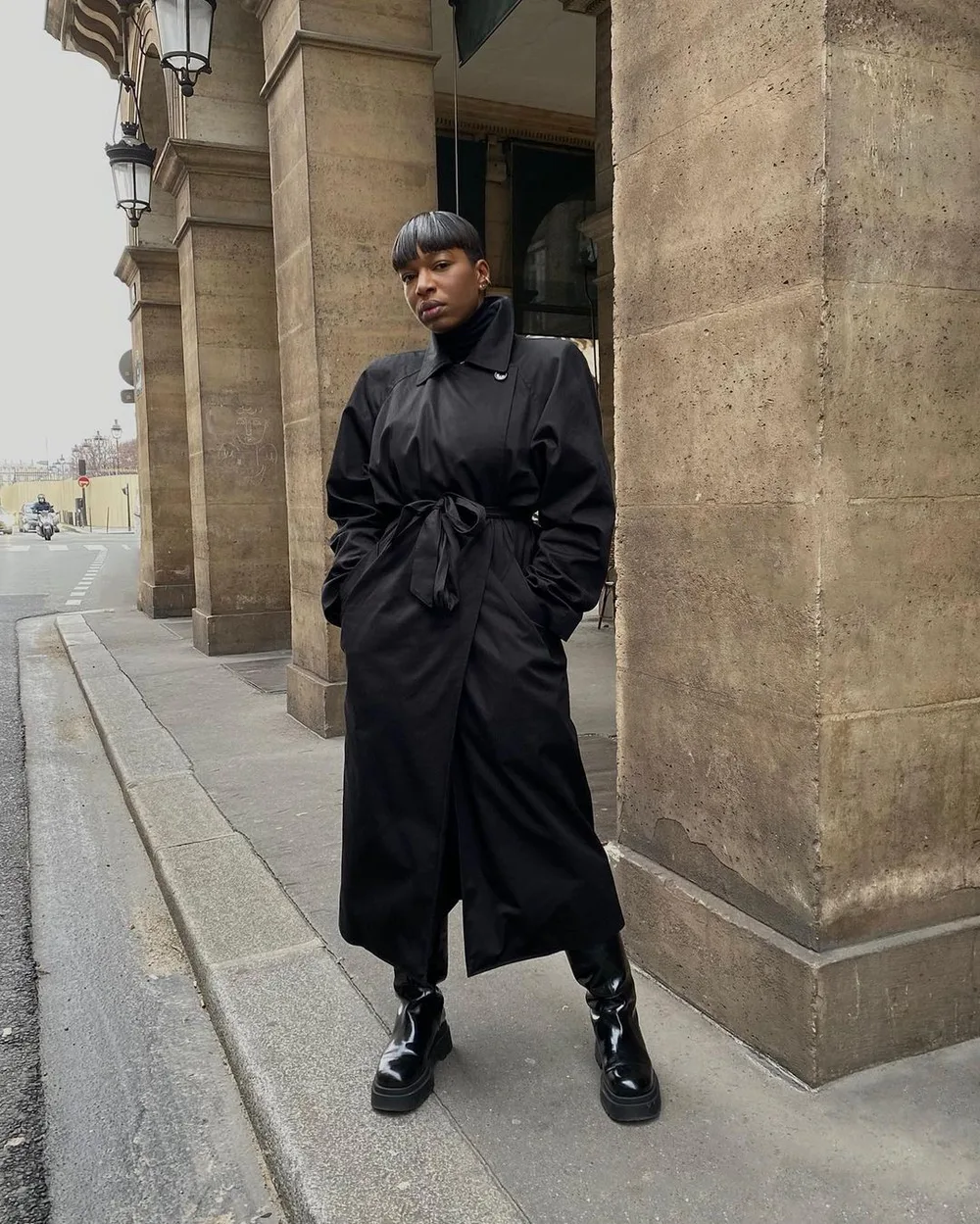 Trench noir long ceinturé porté avec pantalon noir et bottes épaisses