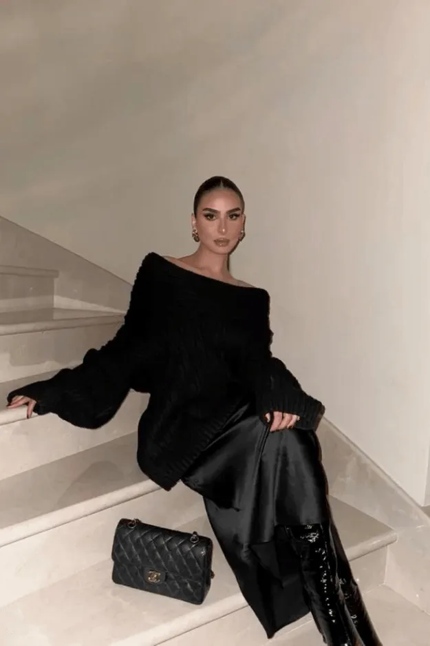 Pull noir aux épaules dénudées avec jupe satinée sombre et bottes hautes